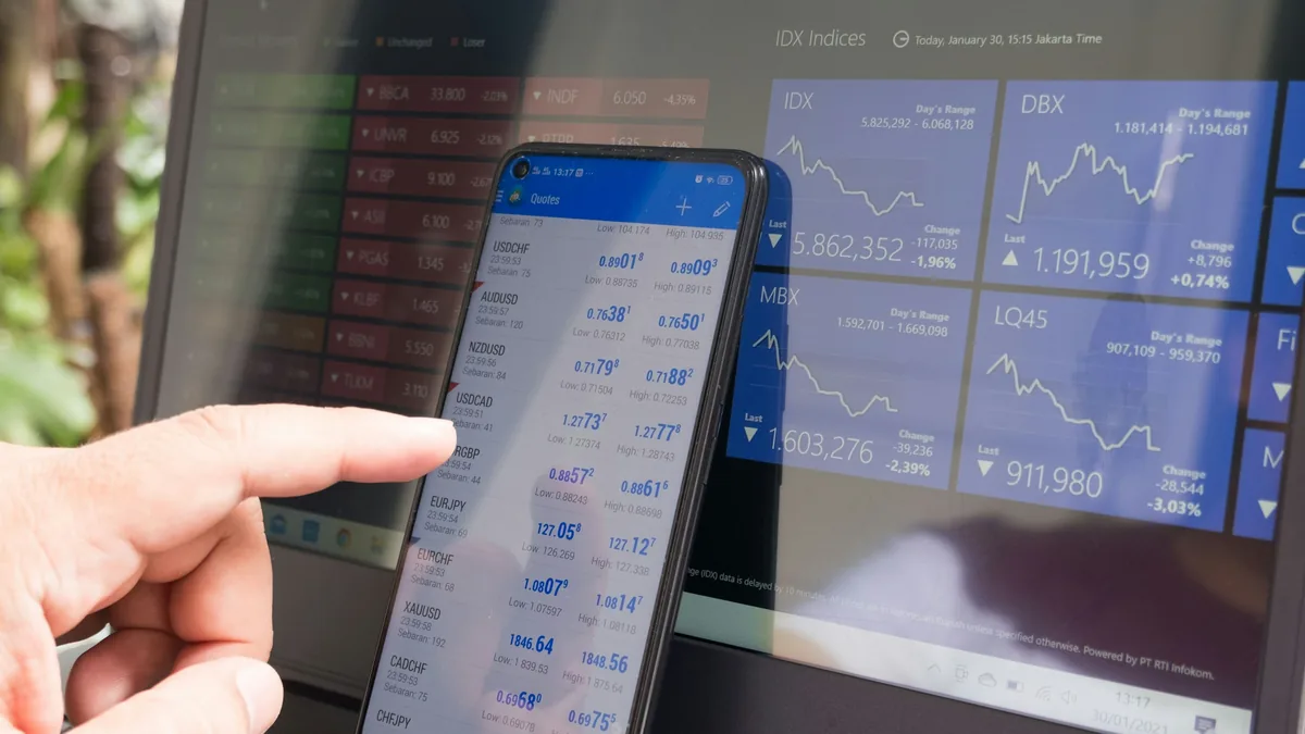 Hand pointing at a smartphone showing currency quotes next to a laptop