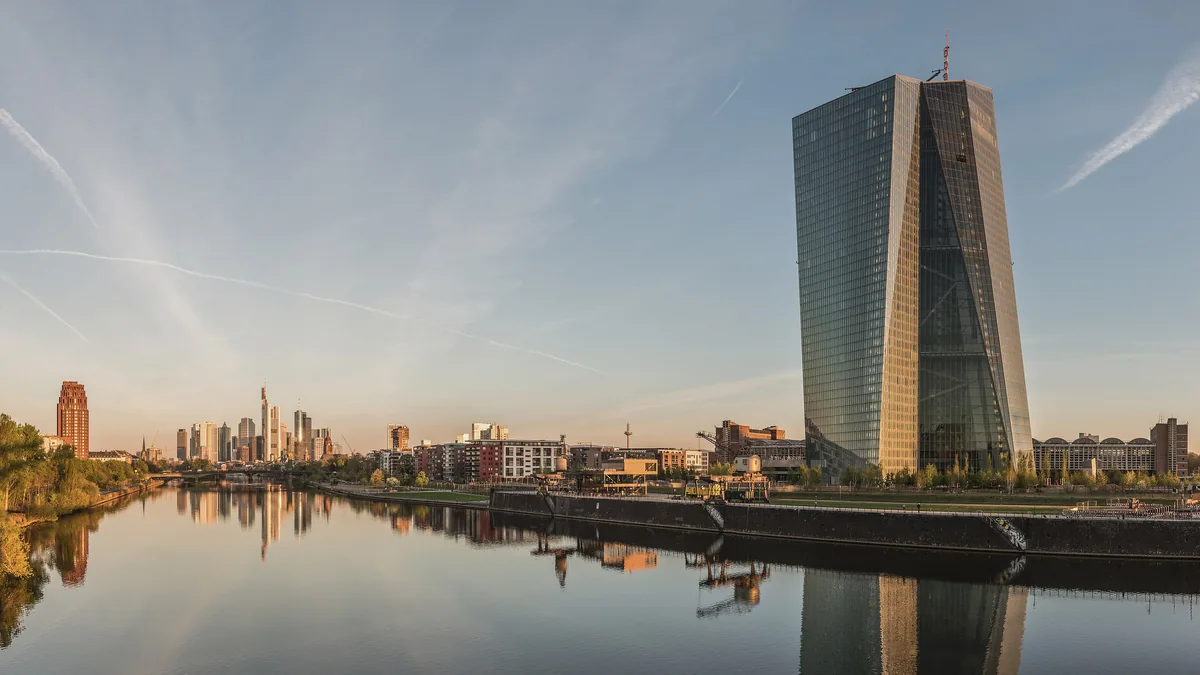 The European Central Bank headquarters, a tall glass tower beside the River Main in Frankfurt at dawn, with the city skyline in the distance