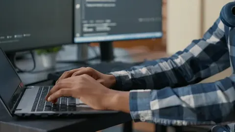 Closeup of programer hands typing machine learning code on laptop keyboard in front of computer screens with programming interface. System engineer writing algorithm for online cloud computing