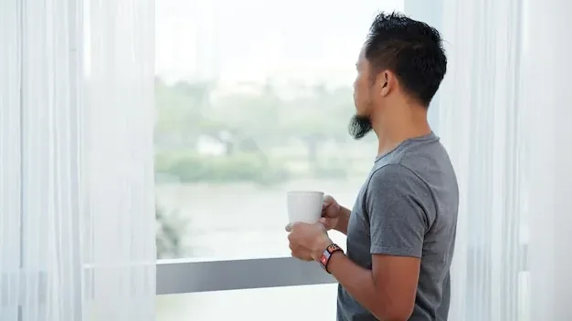 Asian man standing in front of tall window with mug and looking out